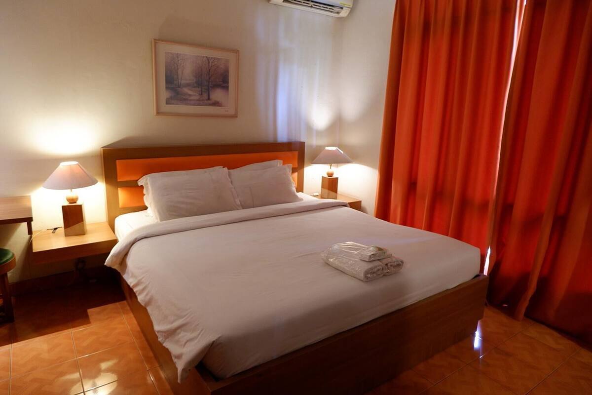 Una cama bien tendida con sábanas blancas, dos lámparas de noche, un cuadro enmarcado en la pared y una cortina roja.