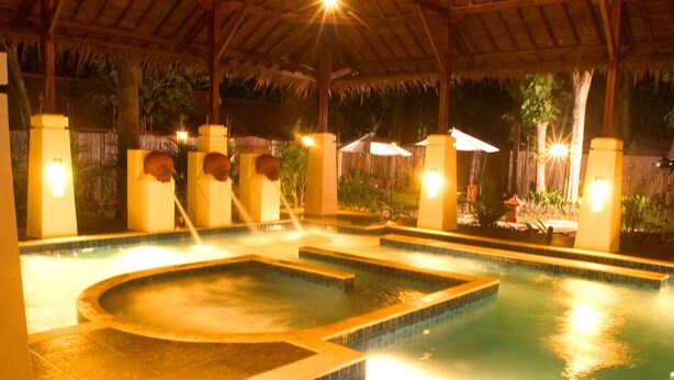A swimming pool with multiple sections, surrounded by pillars and umbrellas, under a thatched roof. - Sanghyang Indah Spa Resort (Anyer)