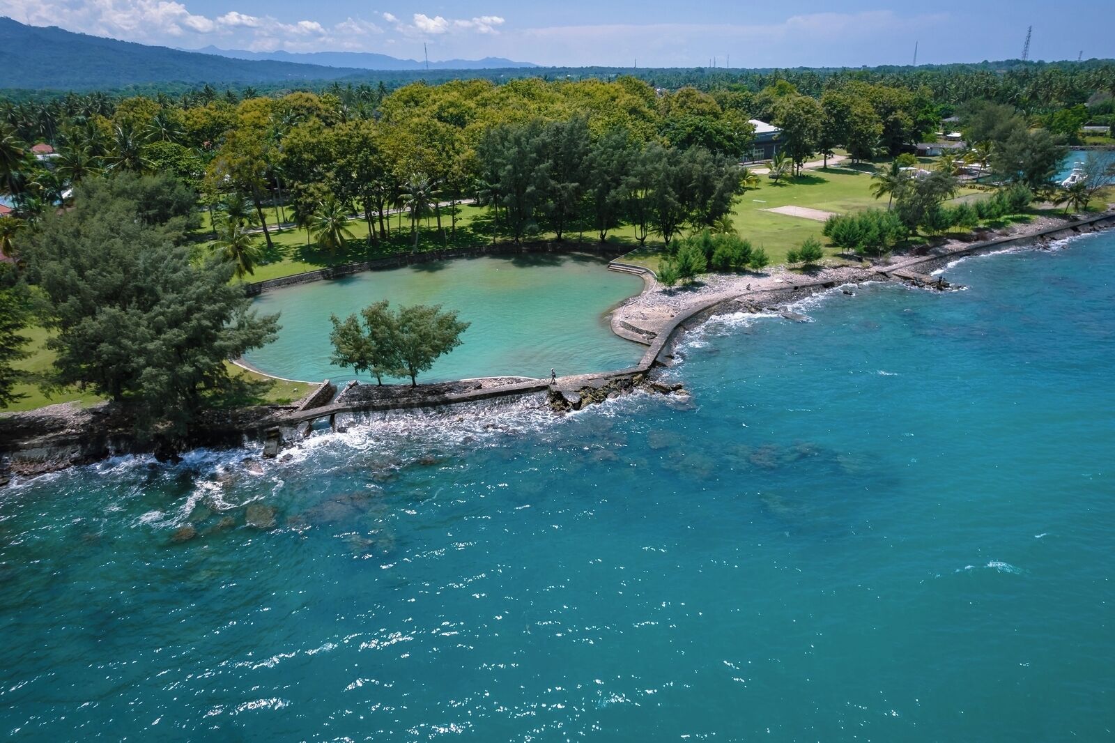 An aerial view of a coastal area with a clear blue sea, a small island with a rocky shoreline, and a golf course in the background.