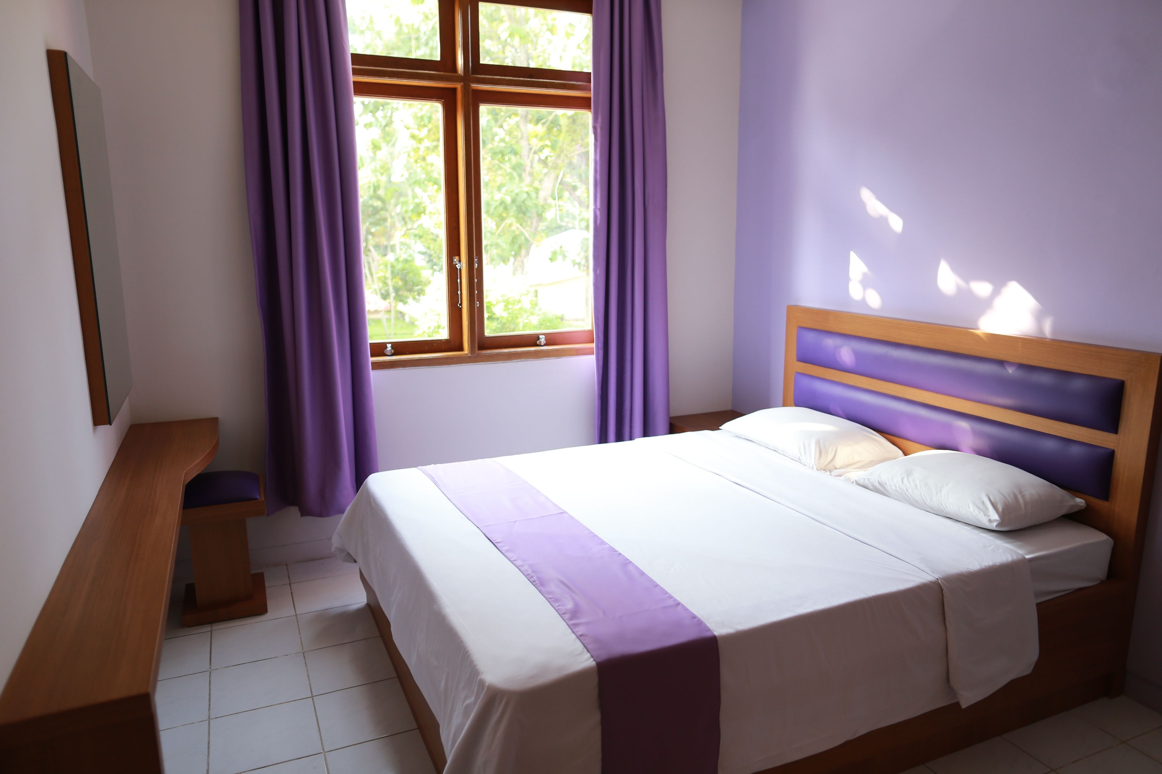 A bedroom with a bed, a wooden bench, a window with purple curtains, and a view of greenery outside.