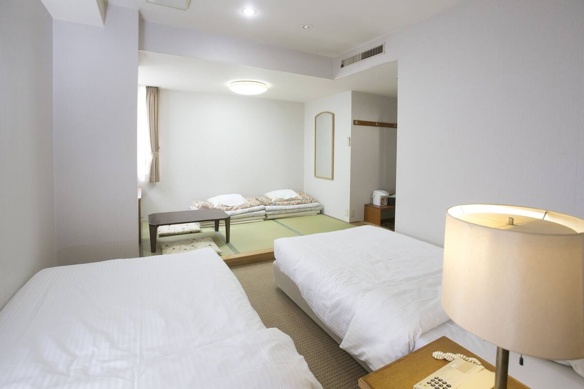 Une chambre d’hôtel avec deux lits, une petite table, une lampe et un futon à l’arrière-plan.