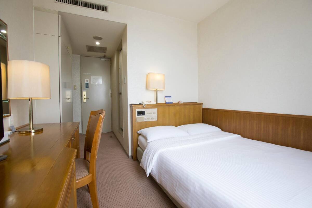 Une chambre d’hôtel avec un bureau en bois, deux chaises, un lit avec du linge de lit blanc et deux lampes de chevet.