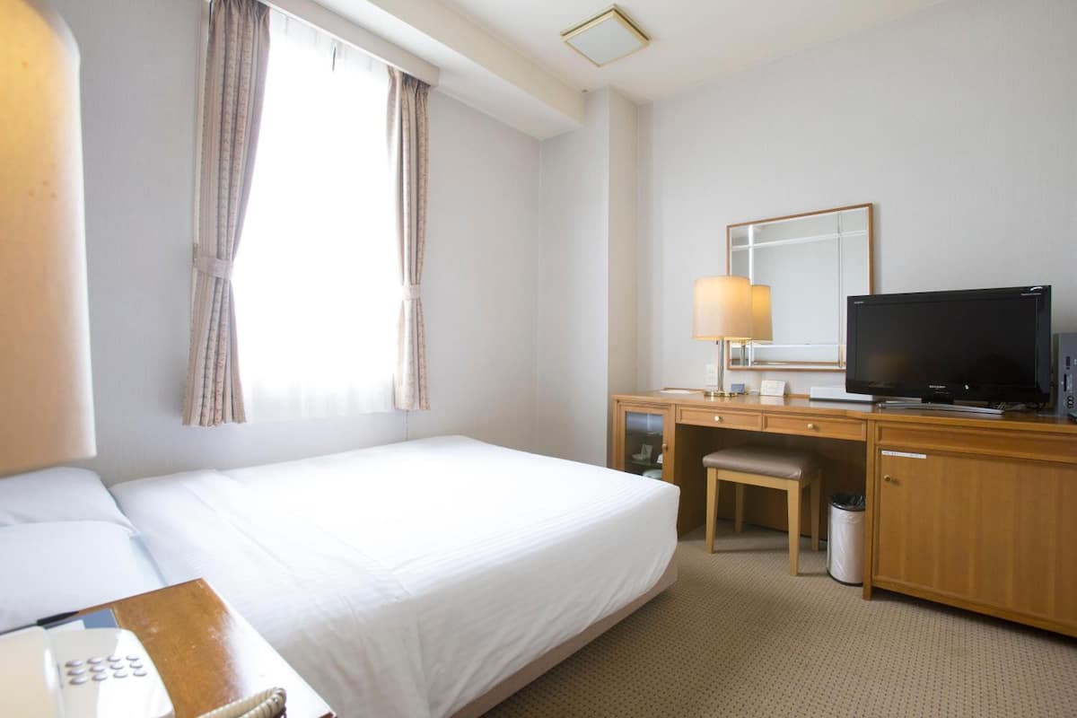Une chambre d’hôtel avec un lit, un bureau avec un ordinateur, une télévision et une fenêtre avec des rideaux.