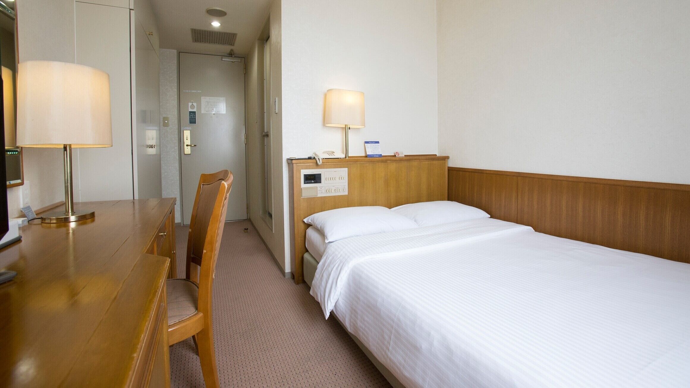Ein Hotelzimmer mit Holzschreibtisch, einem Sessel, einem Bett mit weißen Laken, einem Nachttisch mit Lampe und einer Bedienkonsole an der Wand.