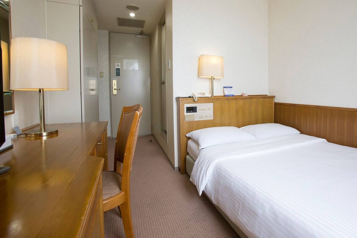 Une chambre d’hôtel avec un bureau en bois, une chaise, un lit avec du linge de lit blanc, une table de chevet avec une lampe et un panneau de commande au mur.
