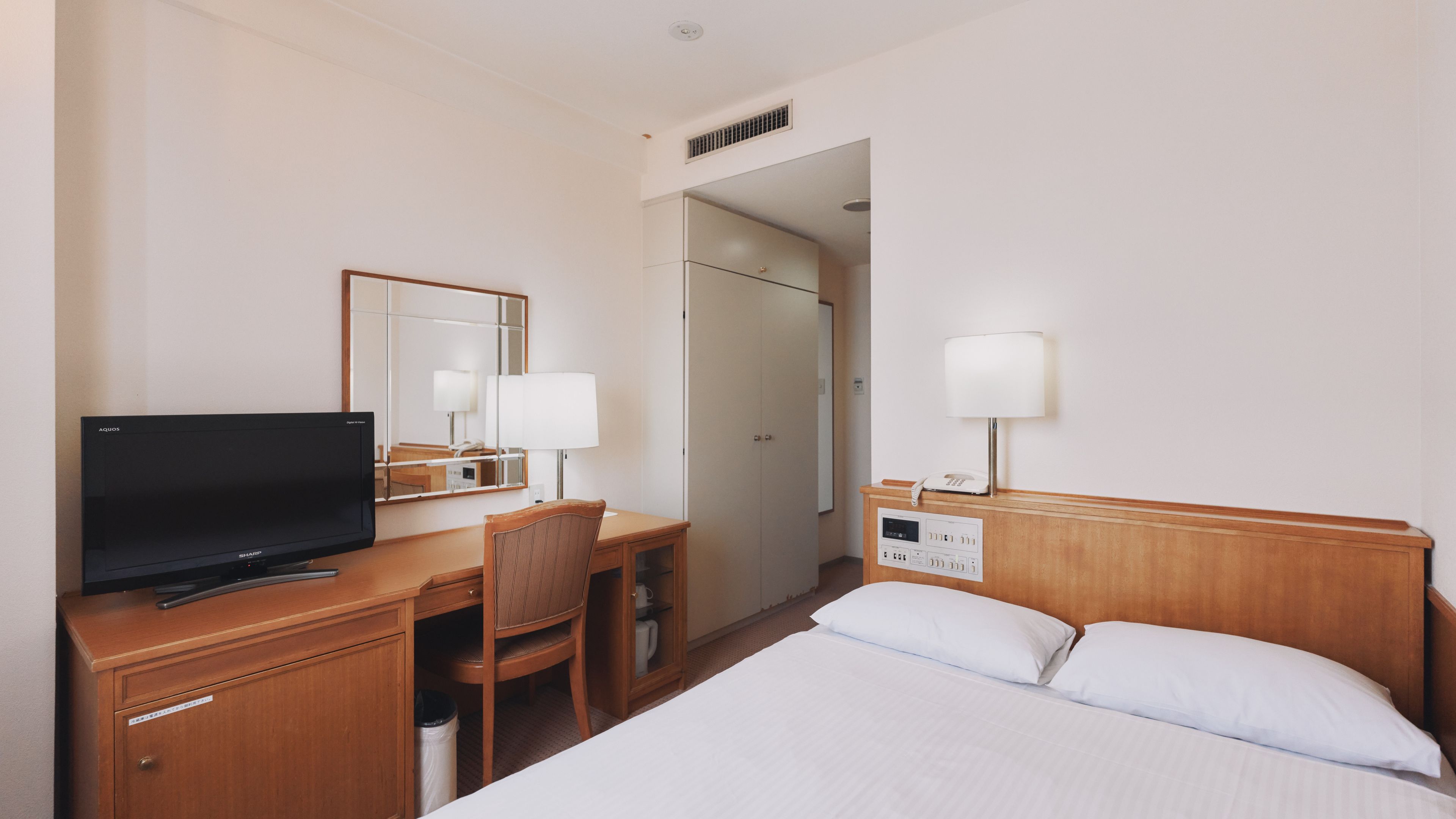 A hotel room with a bed, a desk with a television, a chair, a mirror, a bedside table with a lamp, and a phone.
