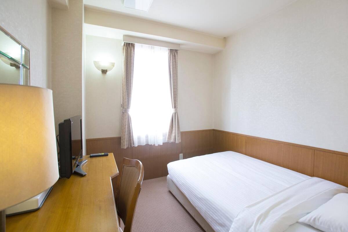 Une chambre d’hôtel avec un lit, un bureau avec une lampe, une chaise et une fenêtre avec des rideaux.