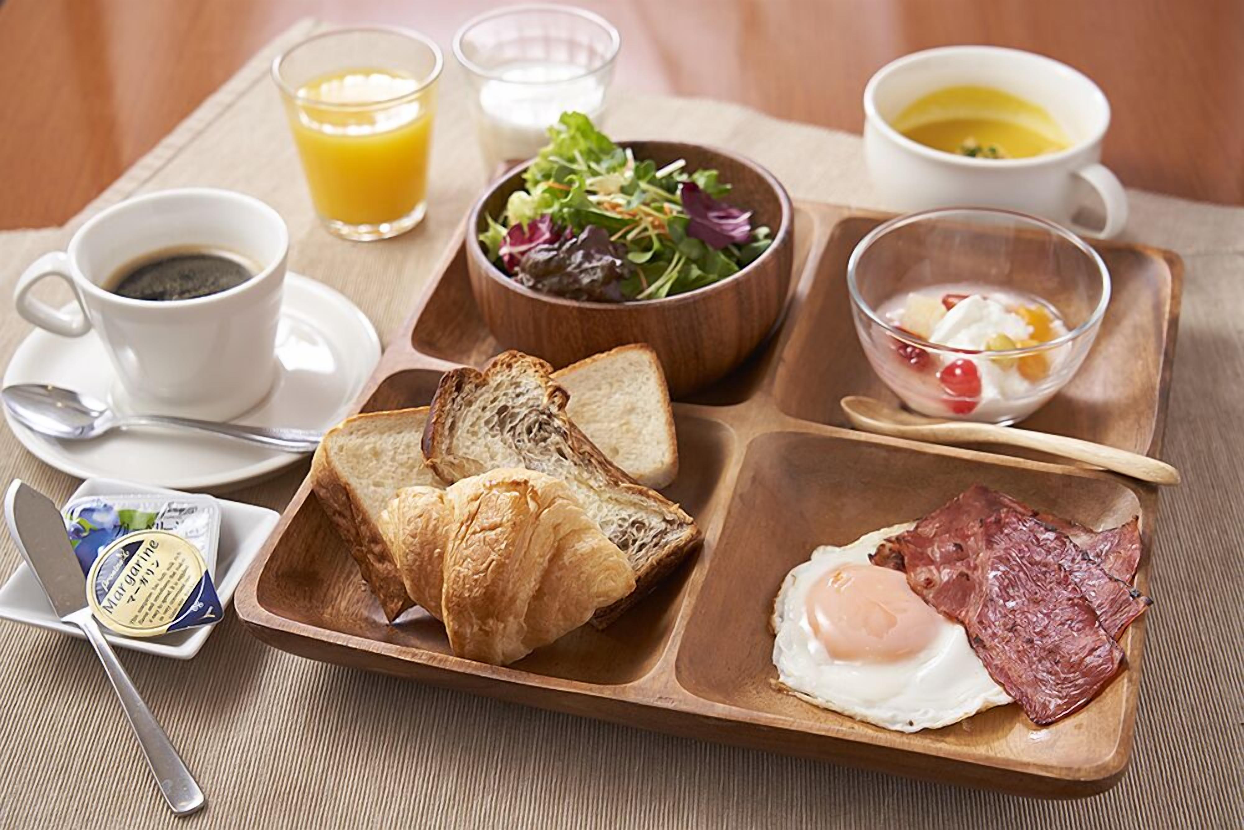 Un plateau de petit-déjeuner avec des toasts, des croissants, du jambon et un œuf, ainsi qu’un bol de salade, un verre de jus d’orange, une tasse de café et un bol de yaourt.