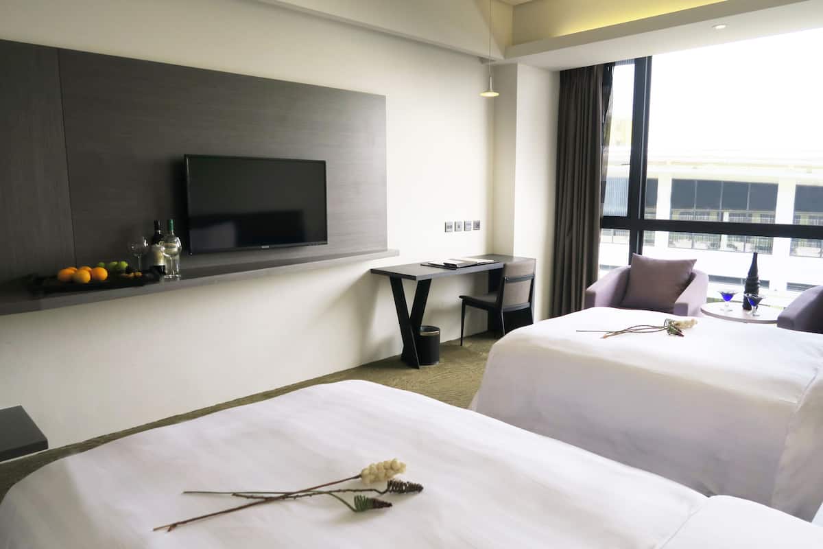 Habitación de hotel con dos camas, un televisor en la pared, una mesita y vistas de la ciudad a través de amplios ventanales.