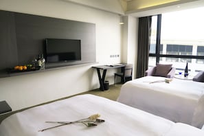 A hotel room with two beds, a TV mounted on the wall, a small table, and a view of the city through large windows.