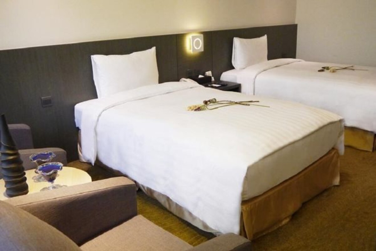 Habitación de hotel con dos camas, una mesita de noche, una lámpara y una pequeña mesa con un florero y dos vasos.