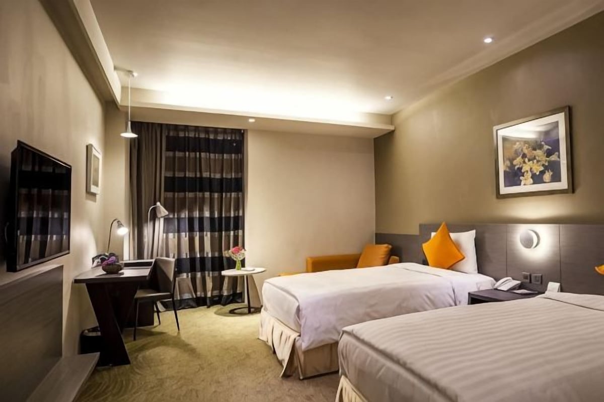 Ett hotellrum med två sängar, ett skrivbord, en stol, ett litet bord med en lampa och en tv.