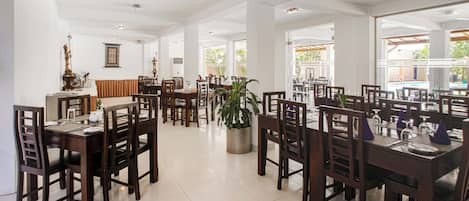 A spacious dining area with multiple tables and chairs, large windows allowing natural light, and a central potted plant.