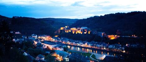 A historic town with a river, surrounded by hills and featuring illuminated buildings and a castle-like structure.
