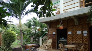 Un patio al aire libre con suelo de hormigón, rodeado de abundante vegetación y palmeras. Hay varias mesas y sillas de madera, además de una mecedora de mimbre. Se ve una planta colgante, y hay carteles en la pared que anuncian alquiler de motocicletas.