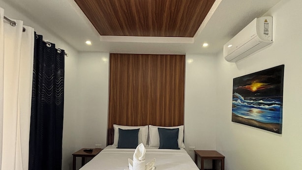Habitación de hotel con techo de madera, cama con sábanas blancas, dos almohadas, un cuadro de un atardecer y un equipo de aire acondicionado.