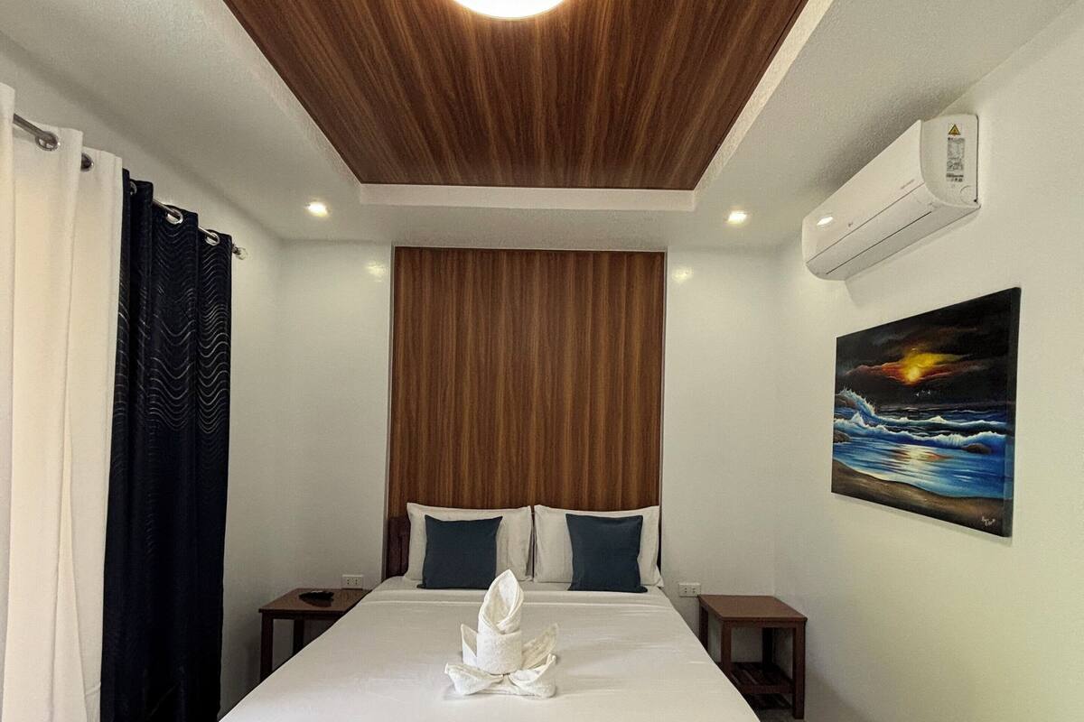 Habitación de hotel con techo de madera, cama con sábanas blancas, dos almohadas, un cuadro de un atardecer y un equipo de aire acondicionado.