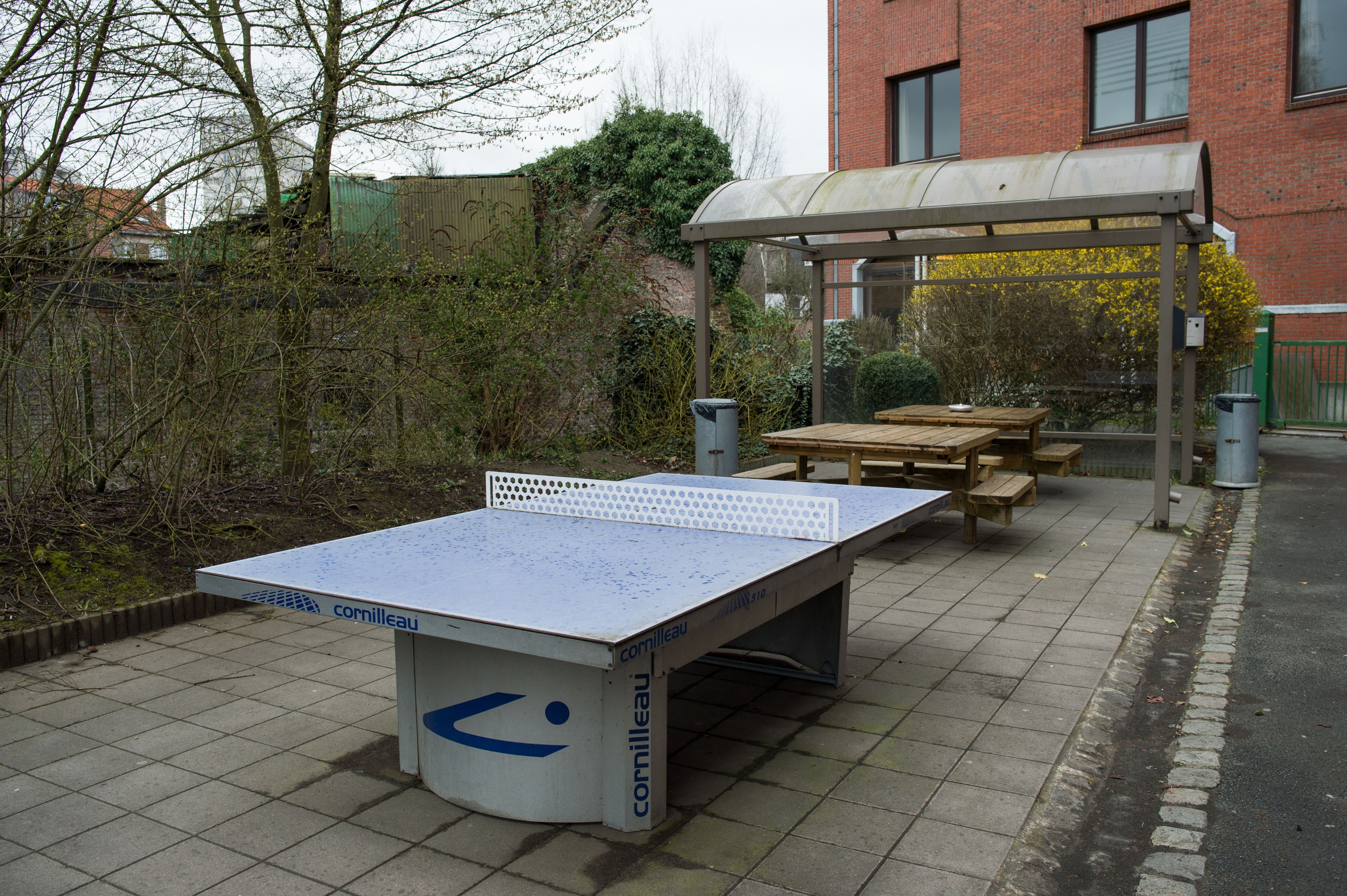 Mesa de ping pong con un toldo, bancos y un cubo de basura en un espacio al aire libre con un edificio de ladrillo al fondo.