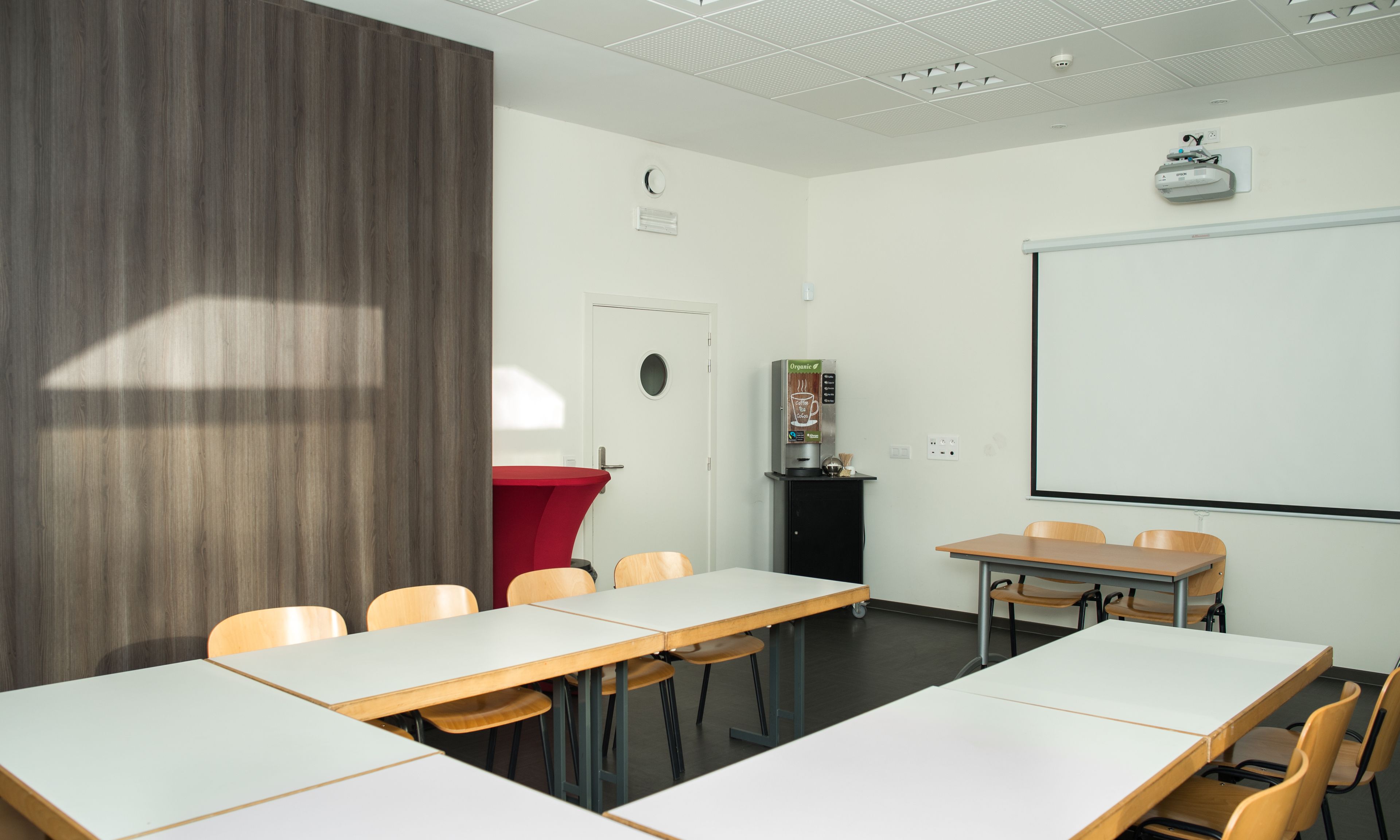 Una sala de conferencias con mesas blancas, sillas de madera, una pantalla de proyector, una máquina de café y una pared de acento roja.