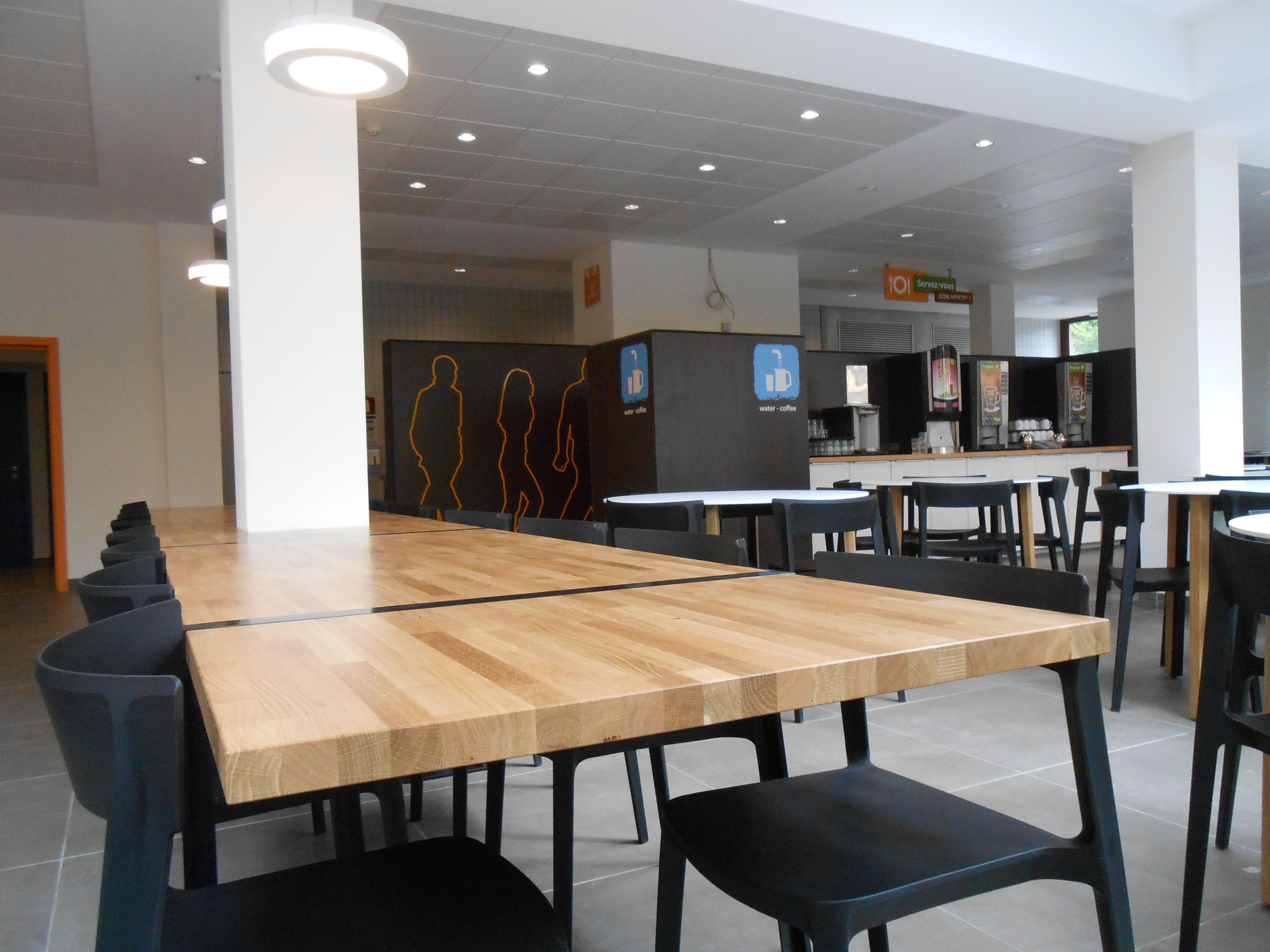Una amplia cafetería con largas mesas de madera y sillas negras, una estación de agua y café, y un mostrador con diversos productos.