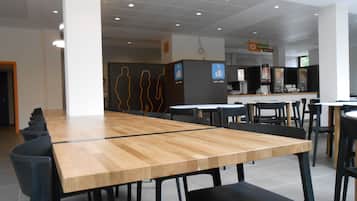 Una amplia cafetería con largas mesas de madera y sillas negras, una estación de agua y café, y un mostrador con diversos productos.