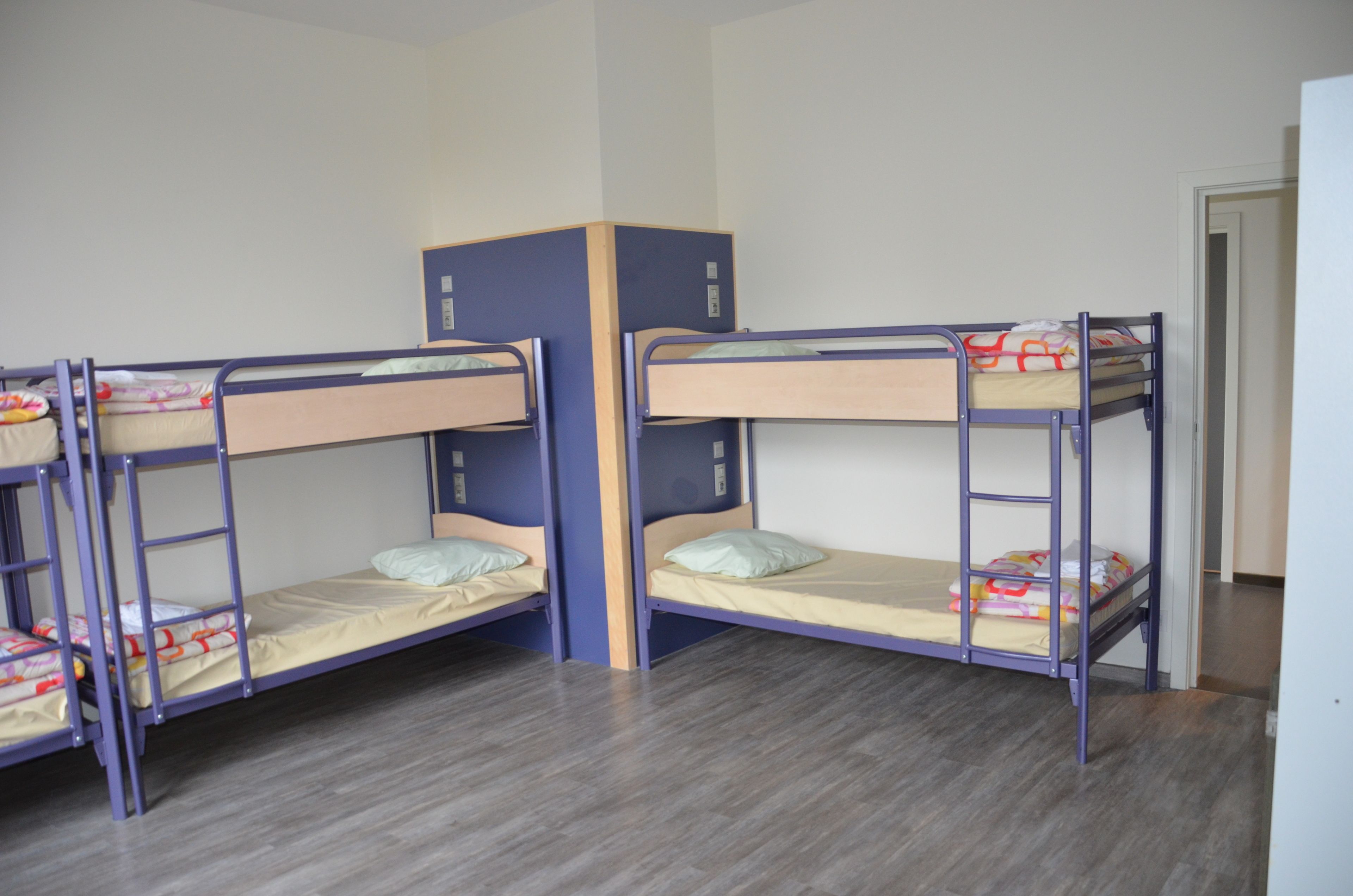 A room with four bunk beds arranged in a corner, wooden flooring, and a door visible in the background.