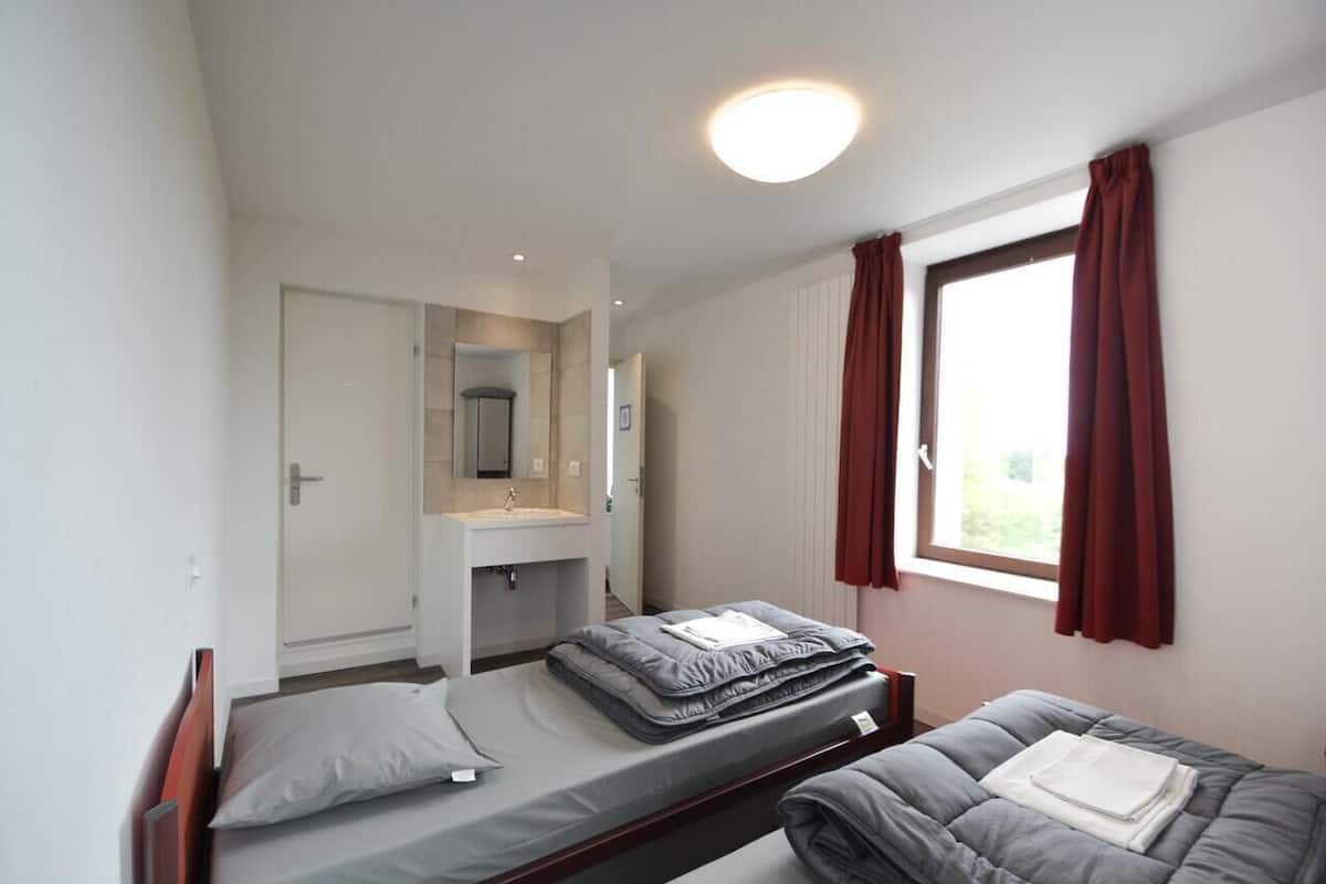A small, clean room with a bed, a bedside table, a mirror, and a window with curtains.