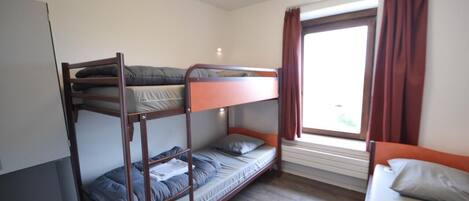 A dormitory room with bunk beds, a window with curtains, and a wooden floor.