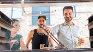 Une personne qui fait griller des aliments à l’extérieur, avec un mur de briques et des fenêtres en arrière-plan.