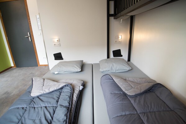 Una habitación con dos camas individuales, cada una con una almohada y una manta. Se observan interruptores de luz en la pared y una puerta.