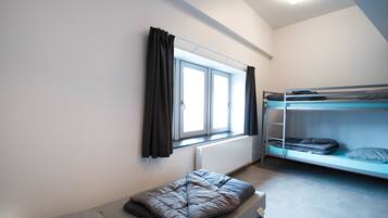 A dormitory room with a single bed, a bunk bed, a window with curtains, and a radiator.
