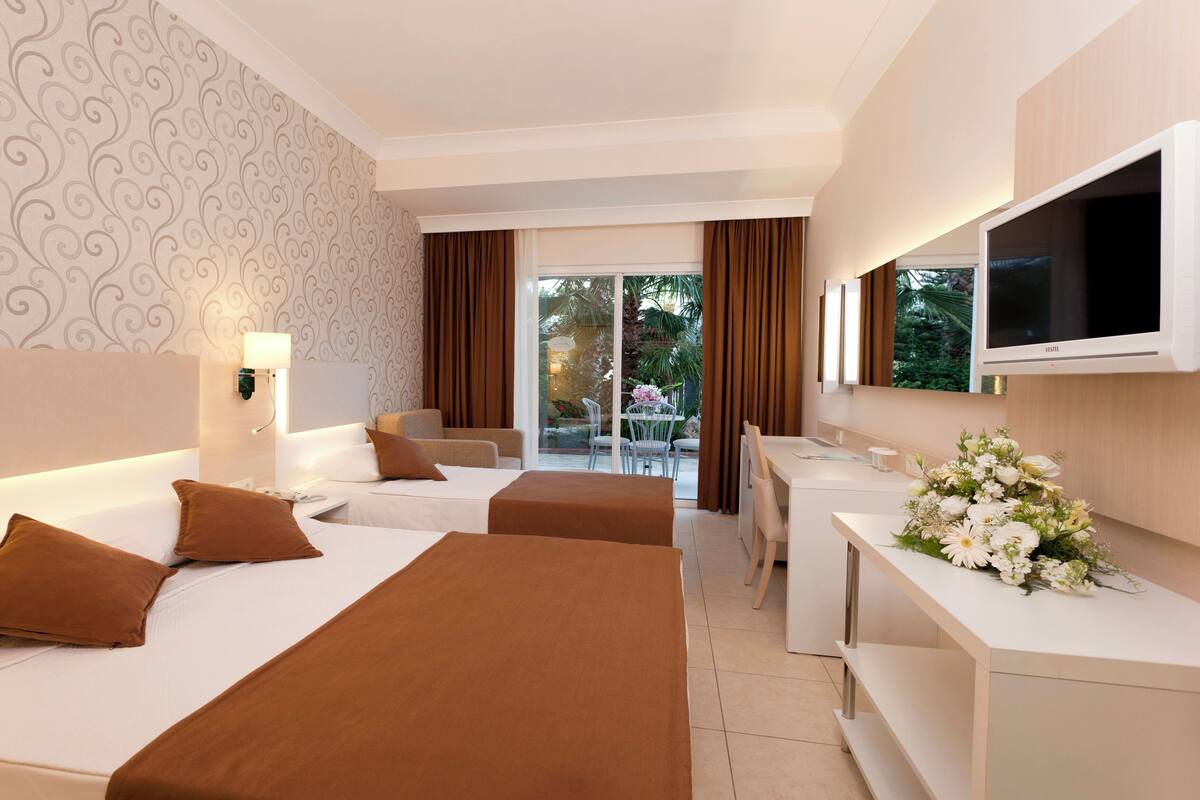 A hotel room with two beds, a desk, a TV, and a balcony with curtains.