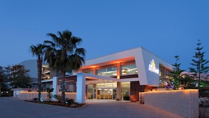 A modern hotel with a glass facade, palm trees, and a well-lit entrance. - Club Mermaid Village  (Alanya)