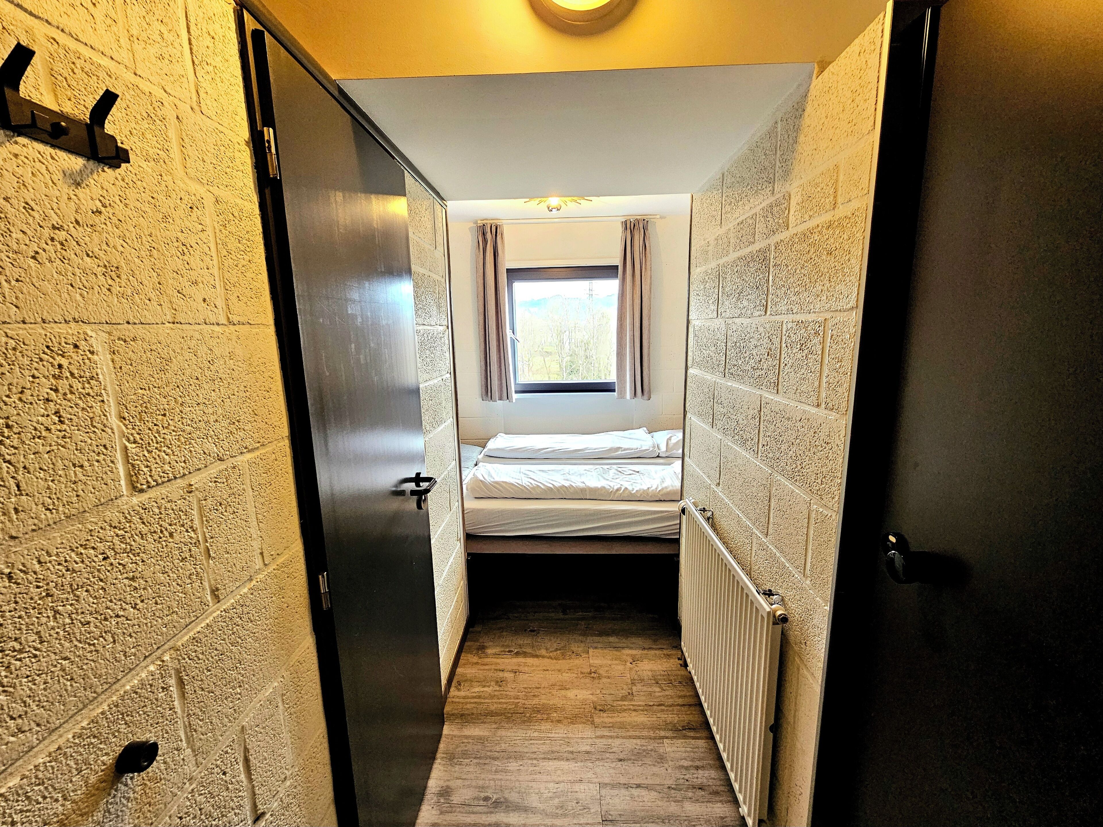 Een smalle gang die leidt naar een kamer met een bed, een raam met gordijnen en een radiator.