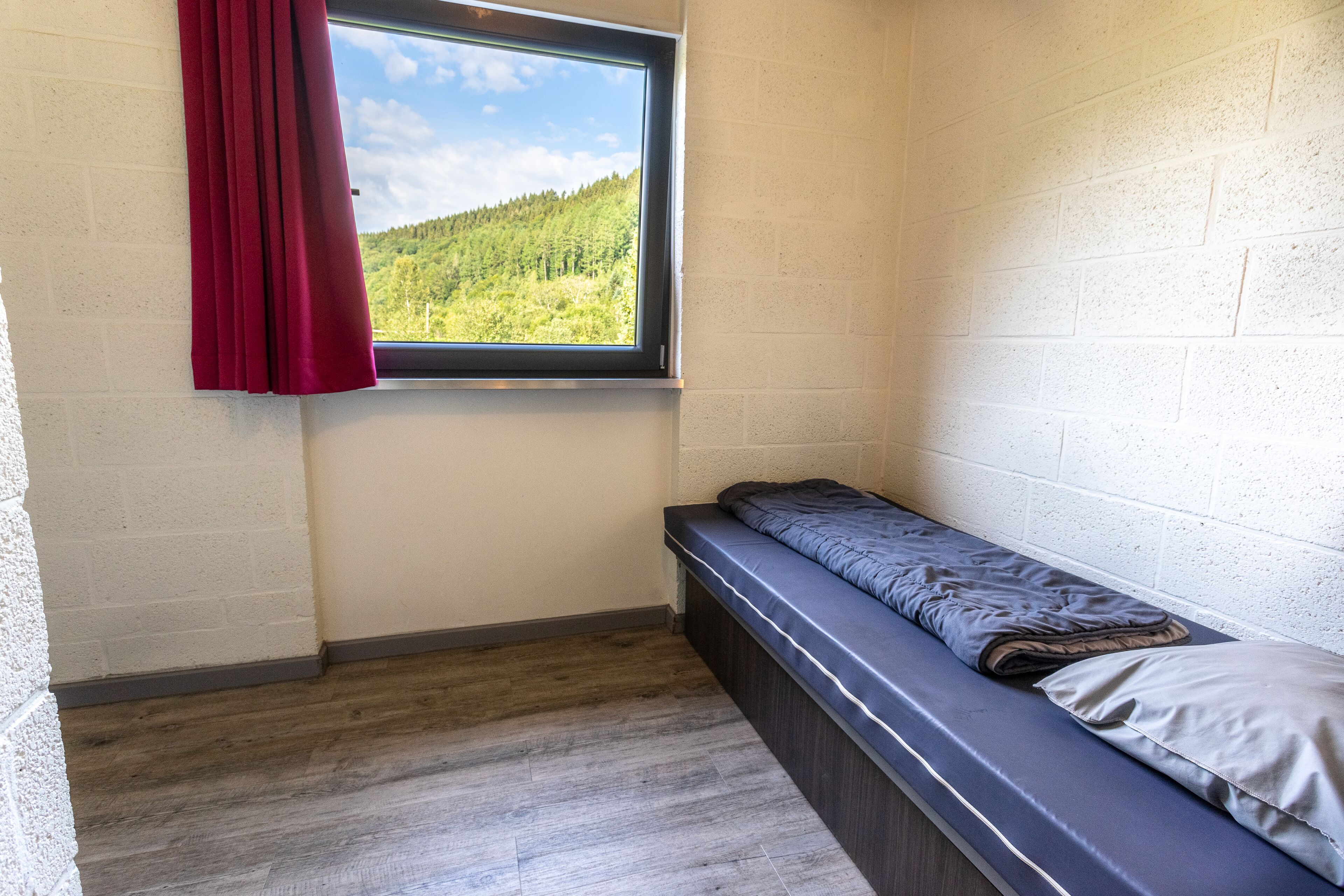 Ein kleines Zimmer mit einem Einzelbett und einem Fenster mit Blick auf eine bewaldete Hügellandschaft.