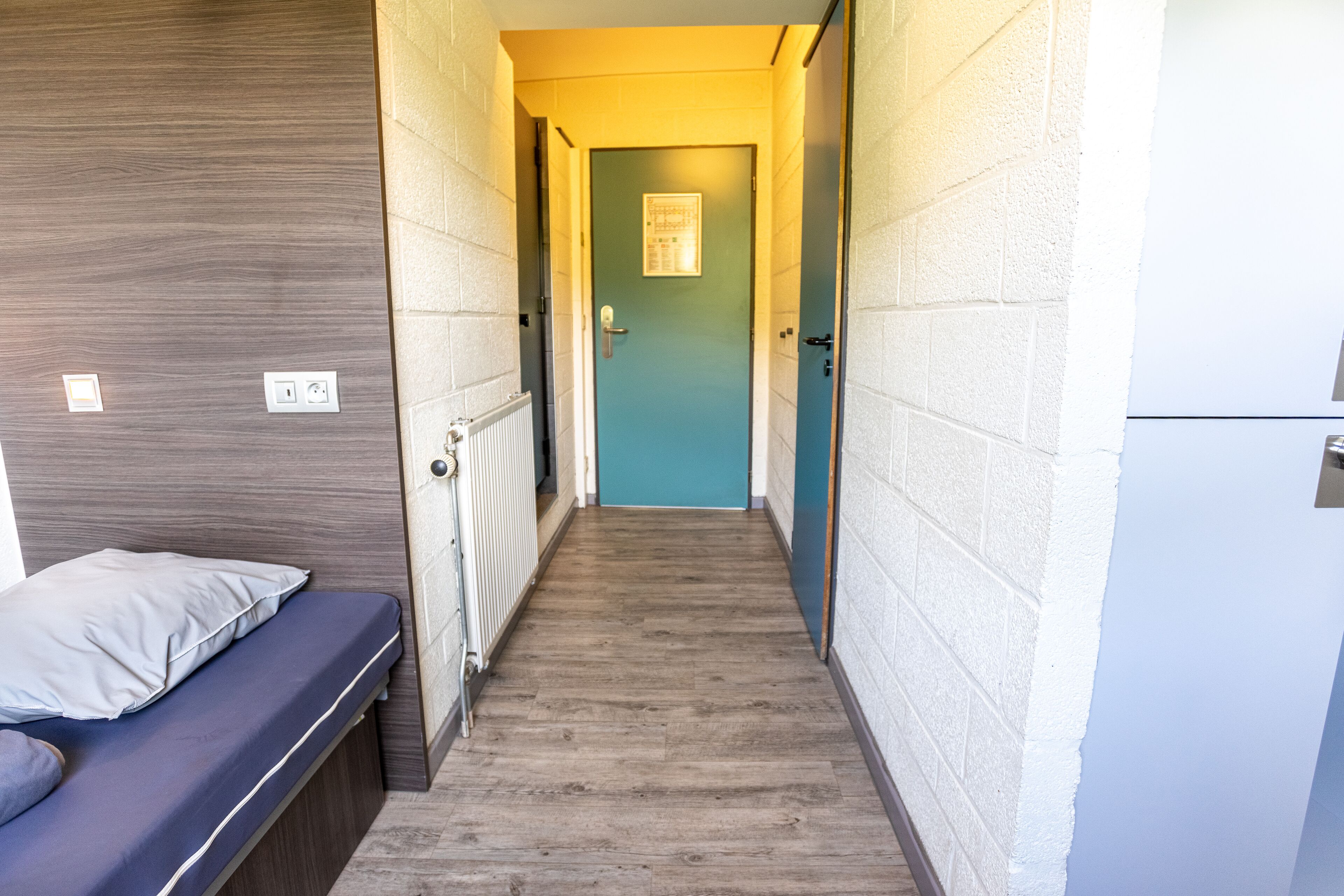 A narrow hallway with a wooden floor, a blue door at the end, and a bed with a pillow on the left.