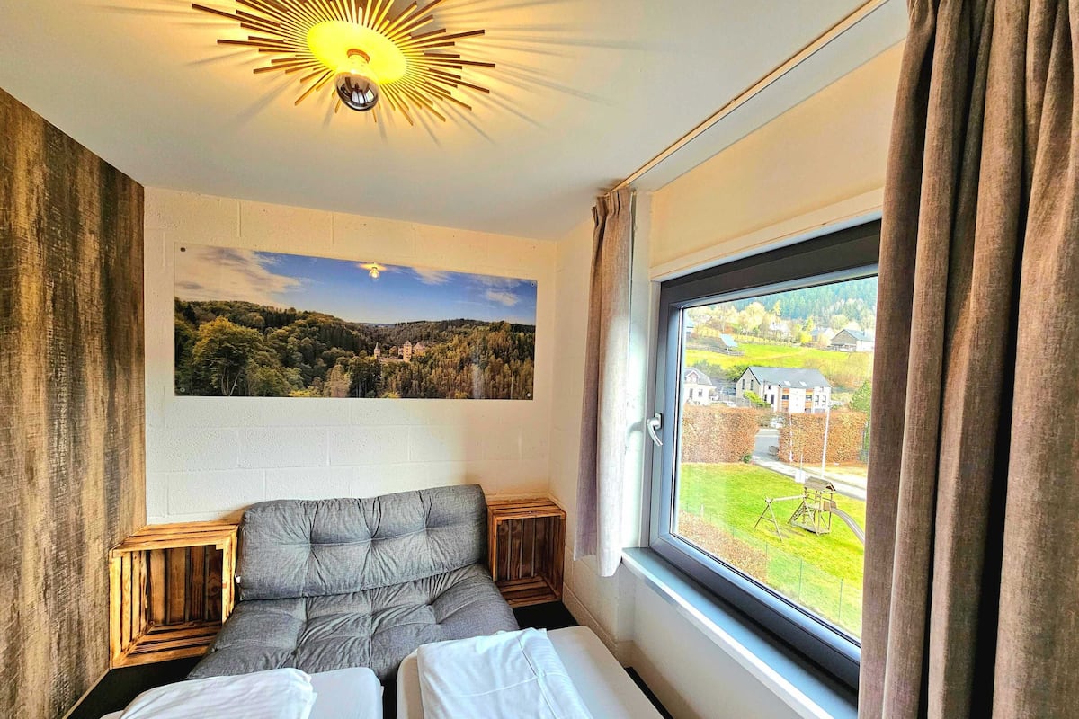 Una habitación de hotel con una cama, un ventanal con vistas al paisaje, un cuadro colgado en la pared y una lámpara de techo original.