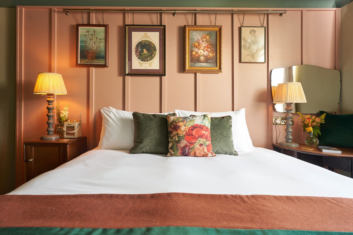 Una cama bien hecha con almohadillas florales, una mesita de noche con lámpara y cuadros en la pared.