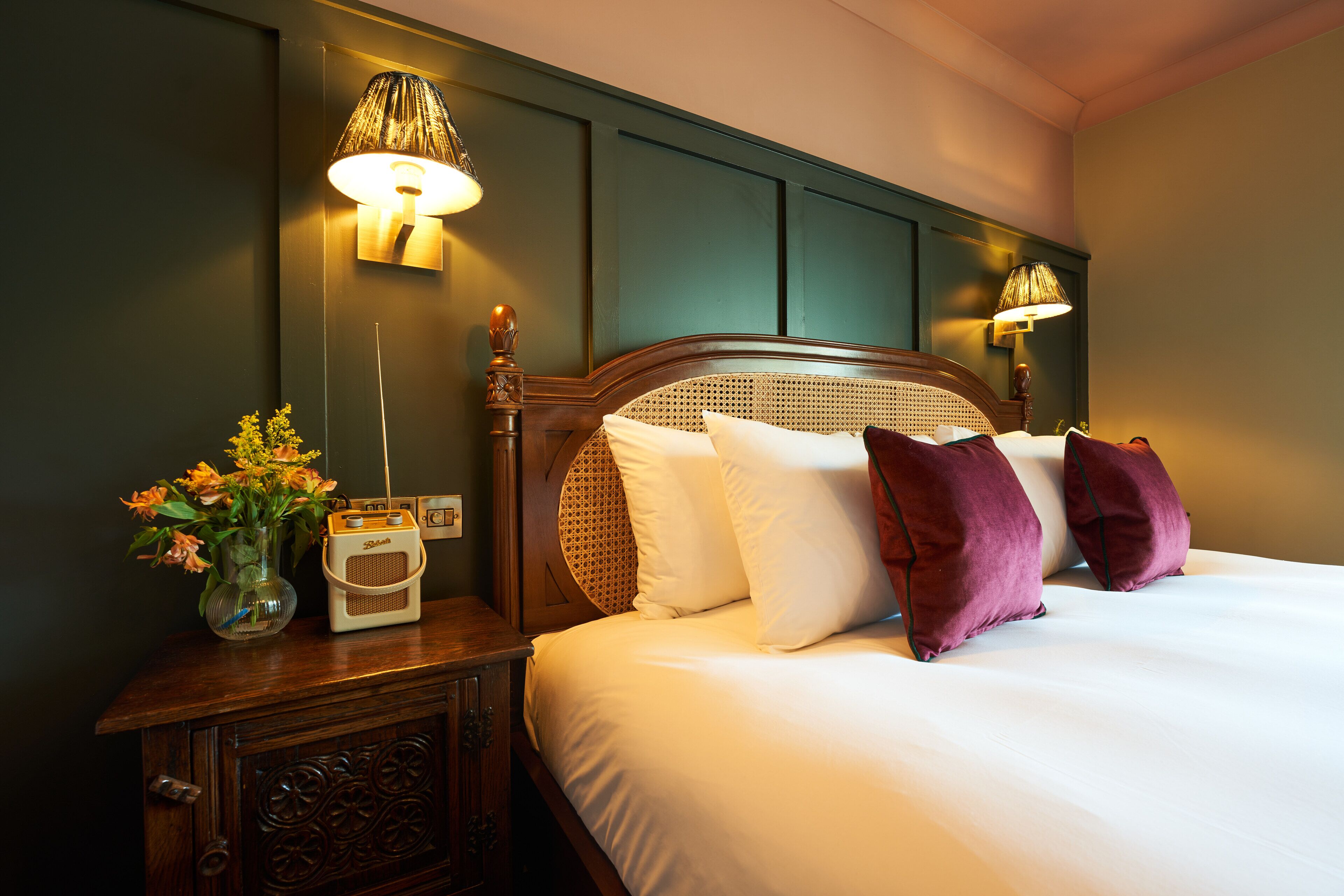 A neatly made bed with white and purple pillows, a wooden nightstand with a radio and flowers, and wall-mounted lamps.