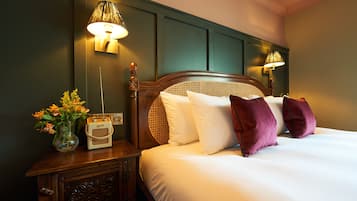 A neatly made bed with white and purple pillows, a wooden nightstand with a radio and flowers, and wall-mounted lamps.