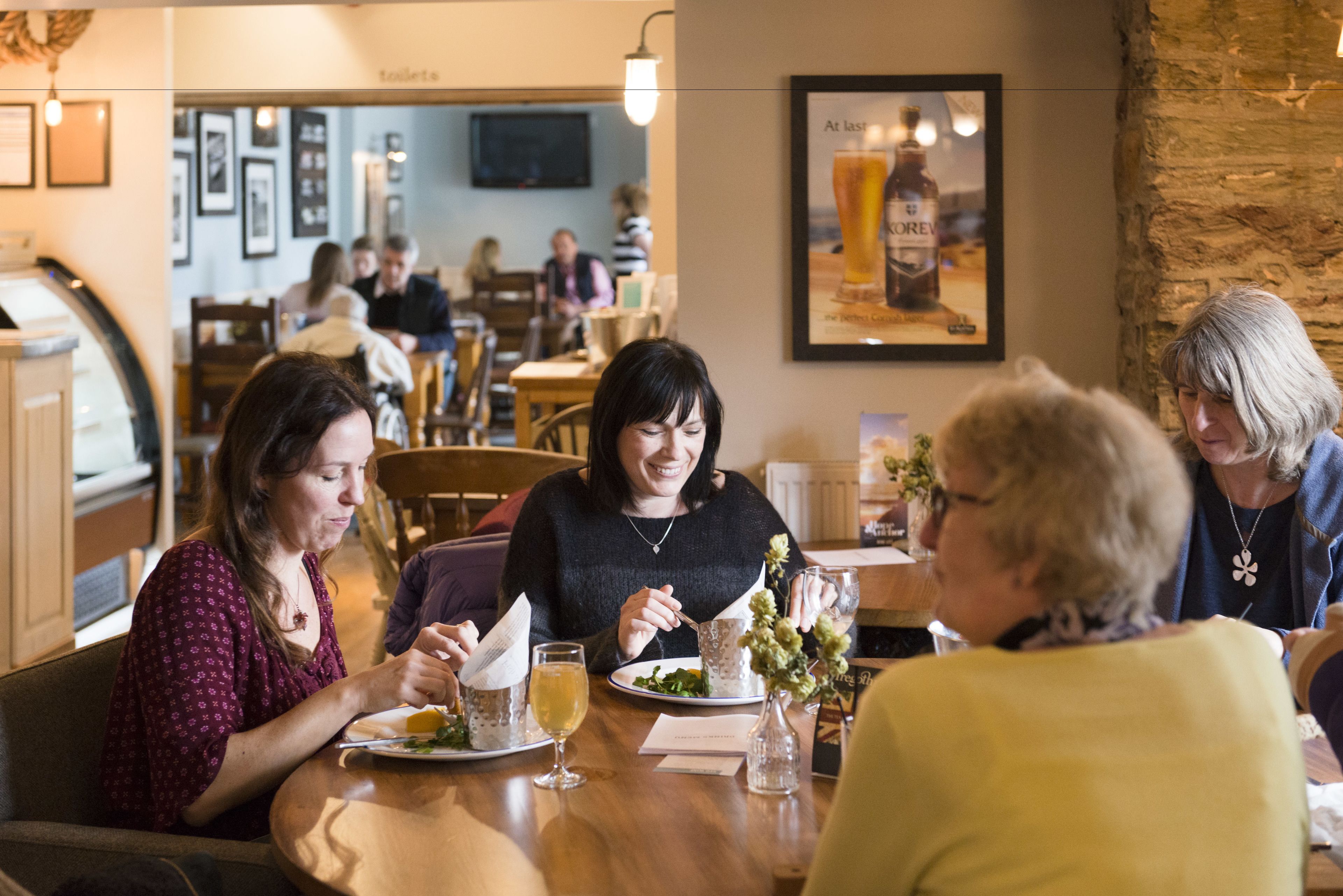 A group of people dining at a restaurant with wooden tables and chairs, a television mounted on the wall, and framed pictures on the walls.