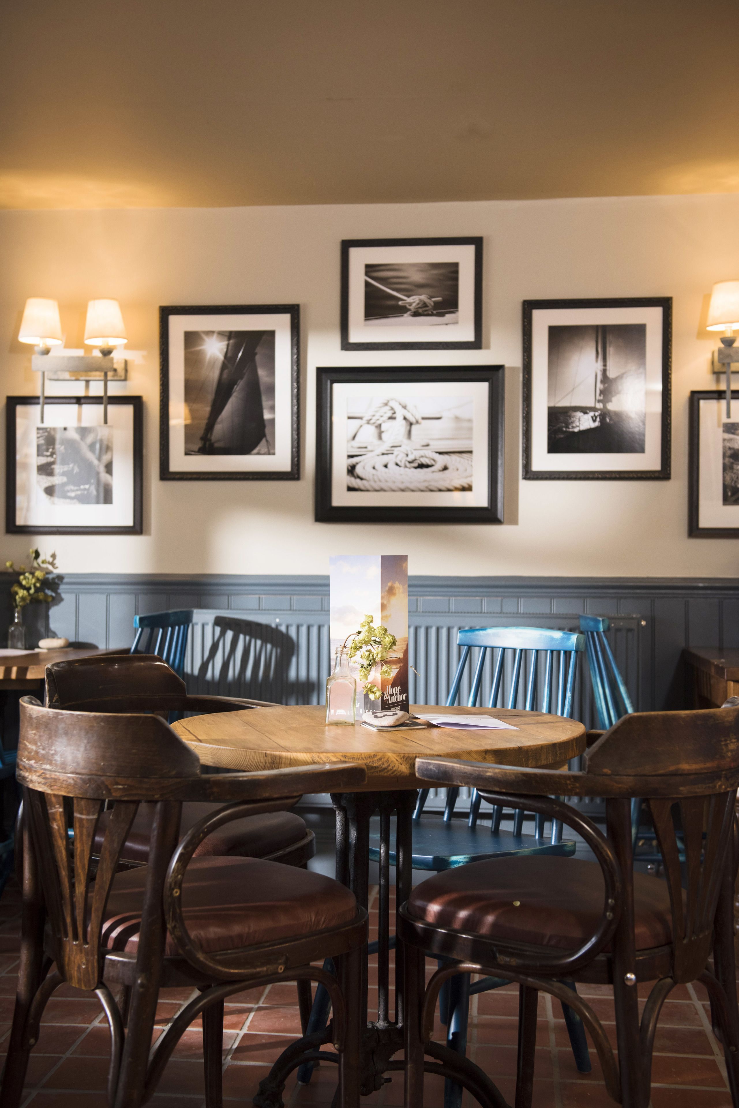 Interior de restaurante con una mesa y sillas de madera, fotografías en blanco y negro en la pared y una silla azul al fondo.