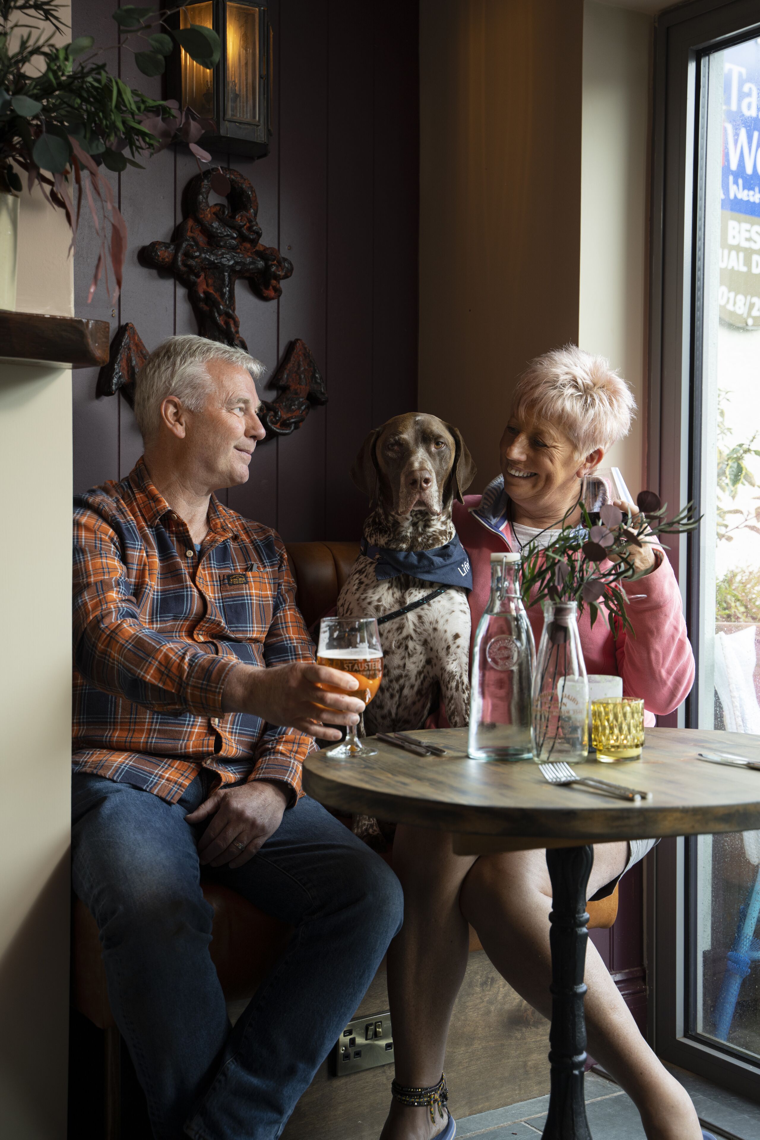 A man and a woman sitting at a table with a dog, holding a glass of beer.