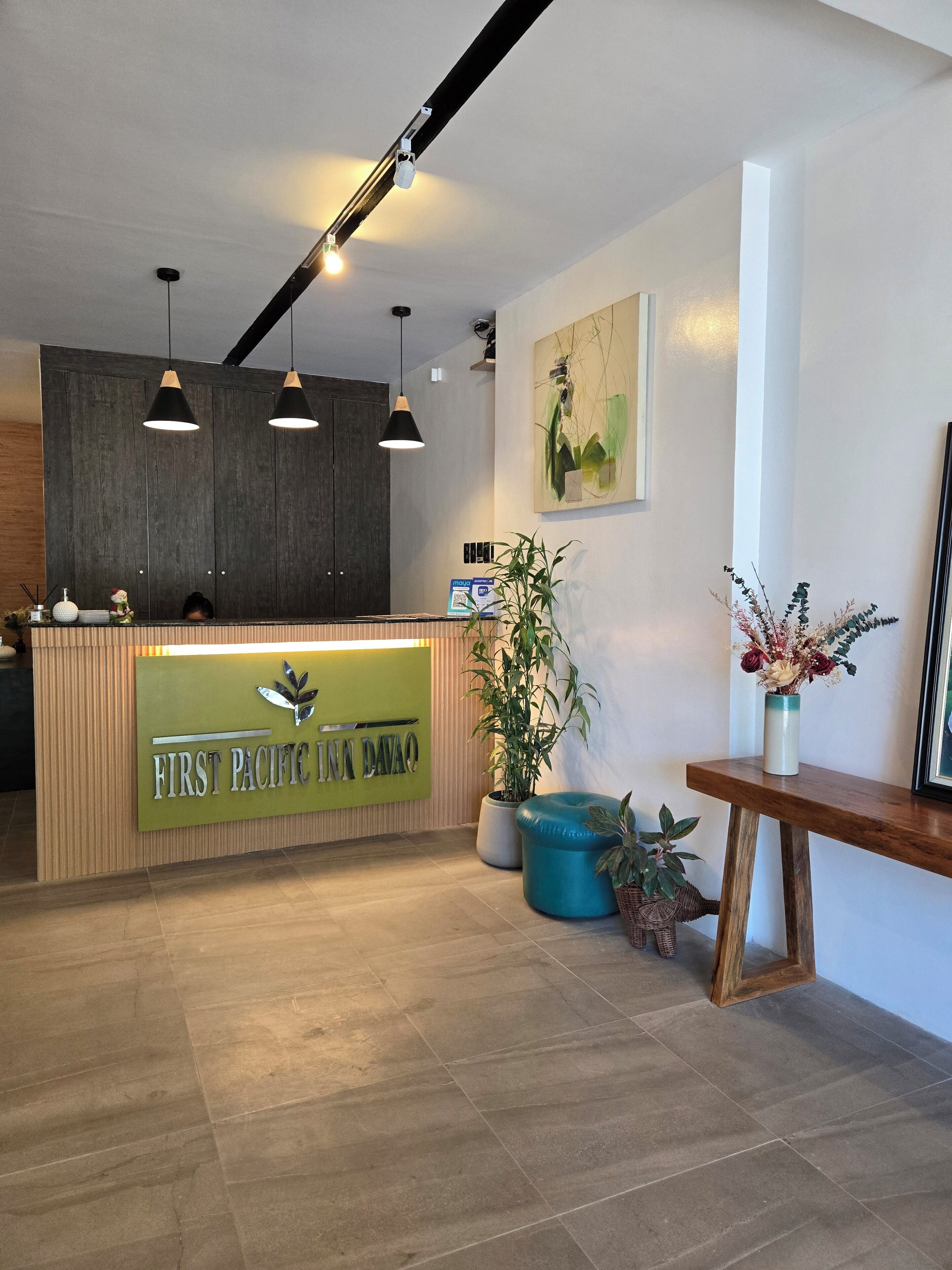 A reception area with a wooden reception desk, a green sign, hanging pendant lights, a large potted plant, and a framed picture on the wall.