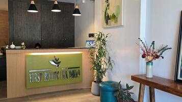 Un área de recepción con un mostrador de madera, un letrero verde, lámparas colgantes, una planta grande en maceta y un cuadro en la pared.
