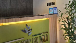 A reception desk with a wooden panel and a green sign that reads "FIRST PACIFIC INN Davao". There are three pendant lights above the desk, and a potted plant is visible to the right. - First Pacific Inn (Davao)