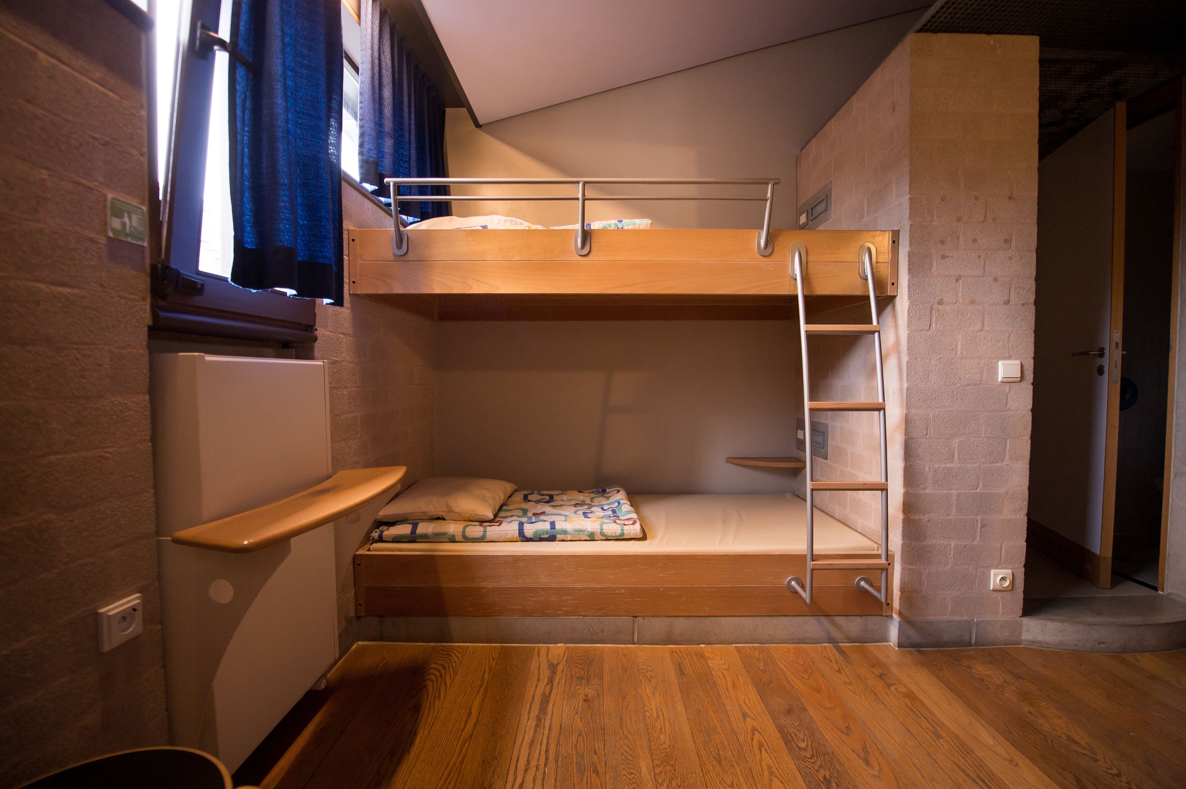 Une chambre avec un lit superposé, un plancher en bois, une petite étagère et une fenêtre avec des rideaux.