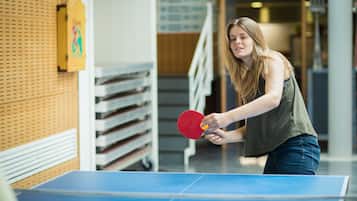 Uma pessoa jogando tênis de mesa em ambiente interno, com uma estante de raquetes e bolas ao fundo.