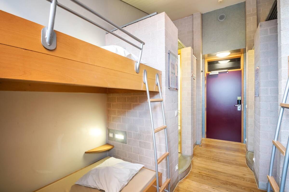 Una habitación pequeña con una cama litera, una mesita de madera, una escalera y una puerta que da a otra habitación.