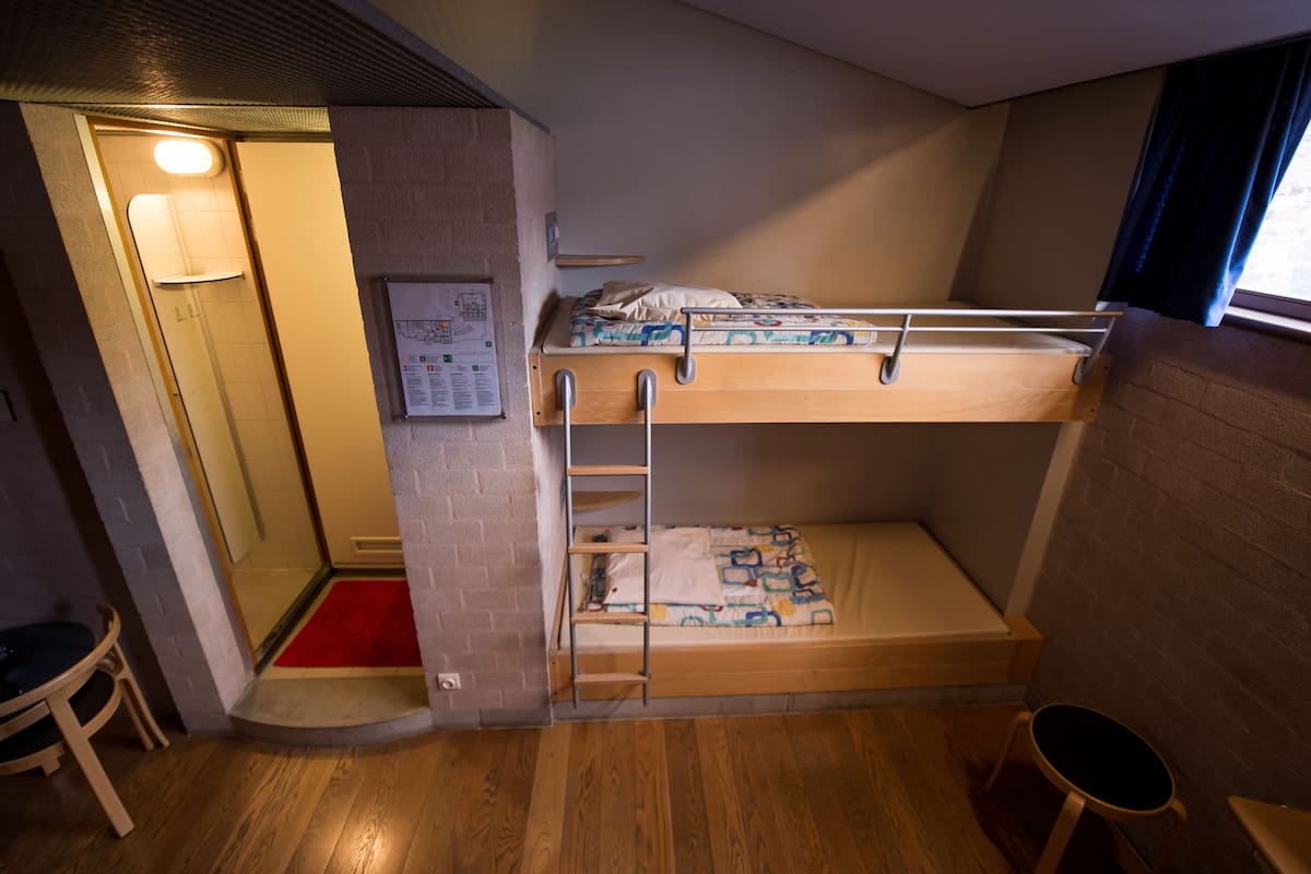 Habitación con literas, piso de madera, un tapete rojo en la entrada, un taburete pequeño y una ventana con cortinas.